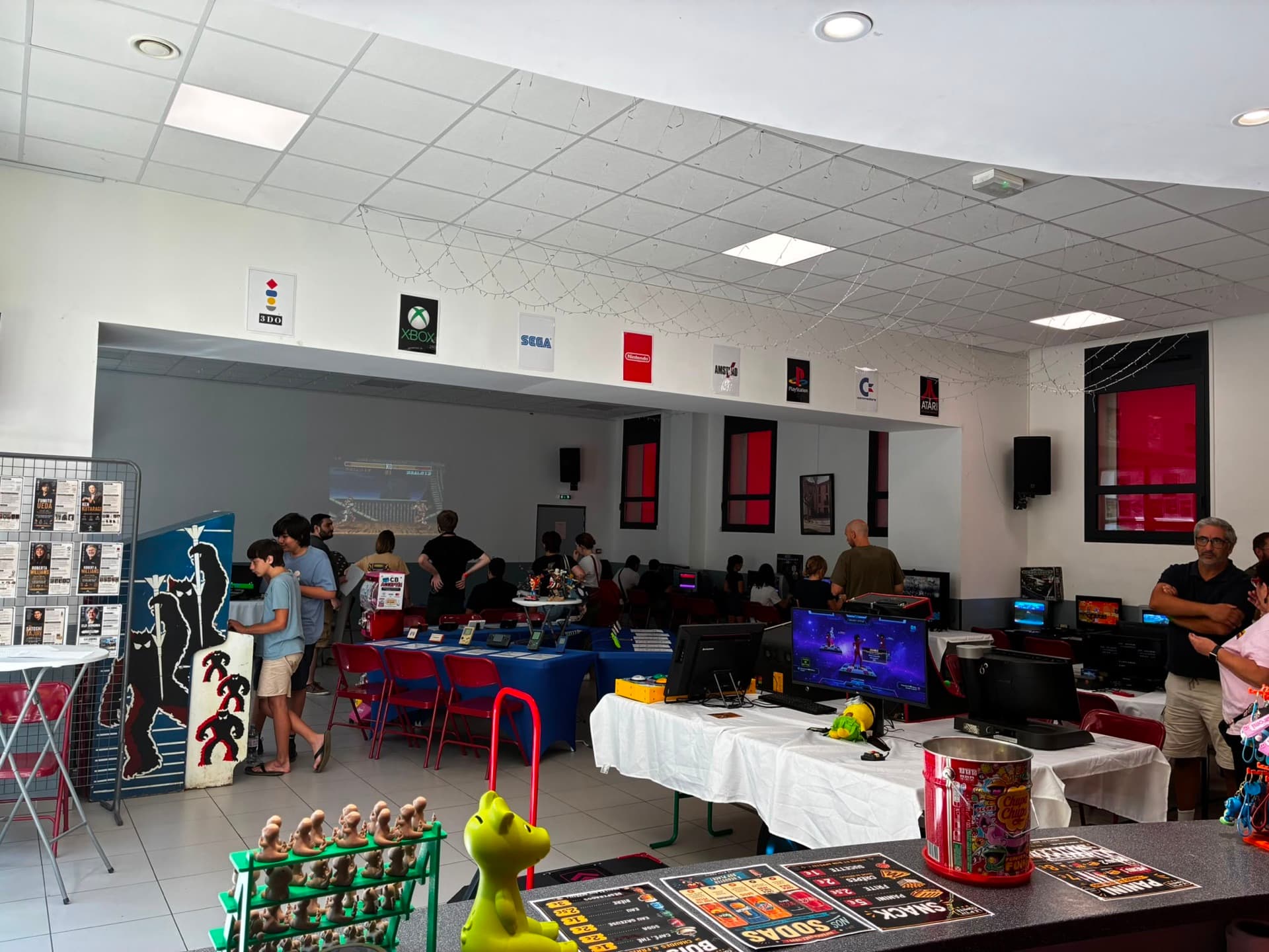 La salle des fêtes de Vingrau pendant le festival Vingrau Level One 2026 : consoles, bornes d'arcade, projection Street Fighter et panneaux SEGA, Nintendo, Atari